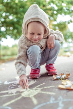 Küçük bir çocuk, yaz şehri, pastel boyalı çizimler, günlük sıcak kıyafetler, kapüşonlu bej kazak. Yaratıcı yaratıcılık ve sanat konsepti. Arka planda sonbahar yaprakları asfalt. Mutluluk duyguları gülümseme ve eğlence.
