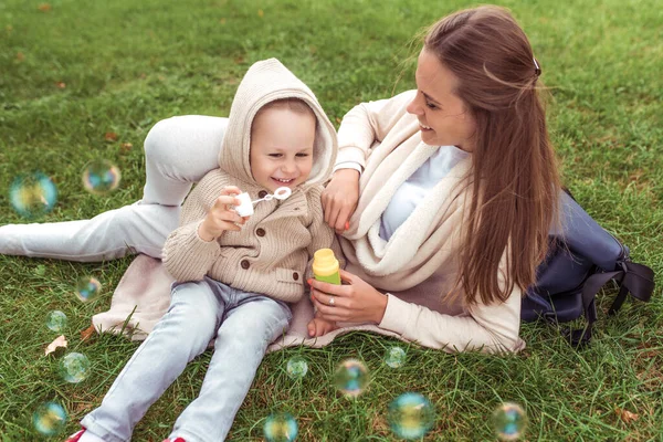 Счастливая молодая семья, женщина-мама, маленький мальчик, надувающий мыльные пузыри, отдых в парке, концепция заботы о детях и любви, поддержка родителей. Фон зеленой травы на газоне. Осень в природе . — стоковое фото