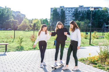 Three girls schoolgirls teens summer on street. Ride on skateboard. Casual clothes, jeans sweaters. Rest after school and lessons. Emotions of comfort, relaxation. Grass lawn building background.