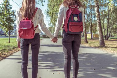 Two girls girlfriends in summer in city, holding each others hands, view from back, autumn day, returning from school and institute, backpacks behind the bags. clipart