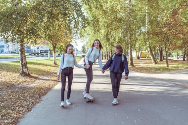Three schoolgirls girls girlfriends in fall in city, ride a skateboard, in summer in park, return from school and college, behind back of bag are backpacks. Casual wear, sweaters, jeans, sneakers.