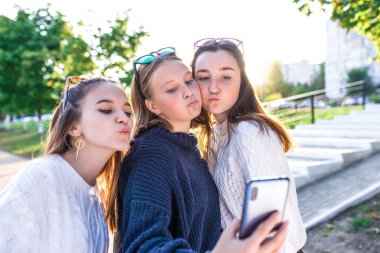 Üç liseli kız, yaz parkı, akıllı telefondan fotoğraf, telefonda selfie. İnternete çevrimiçi video bağlantısı. Ilık örülmüş kazaklar. Okul bittikten sonra yürüyüş eğlenceli duygular..