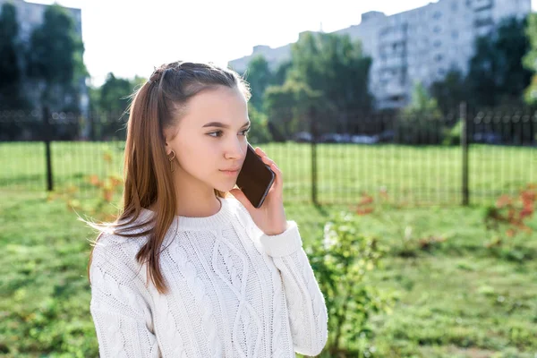 夏の街で白いセーターの10代の女の子。スマートフォンで呼び出し、音声メッセージに耳を傾ける。普段着。コピーテキストのための無料スペース。緑の芝生の建物の背景. — ストック写真