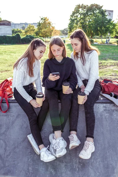 3 Mladé dívky školačky teenageři sledování videa na telefonu. Oblečte si džíny. Emoce relaxační přestávky na vysoké. Nejlepší přítelkyně, relaxace po škole, šálky čaje, tašky, batohy. — Stock fotografie