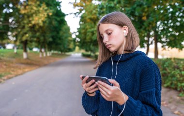 Ciddi bir genç kız öğrenci, yaz parkı, elinde akıllı telefon, video kulaklık izliyor. Okul bittikten sonra parkta dinlen. Öğrenmeyi anlayan duygular, video dersi.