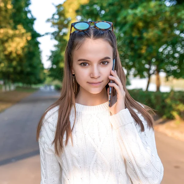 Девочка-подросток 12-15 лет, в белом свитере, летом в городе. Звонки на смартфон, прослушивание голосового сообщения. Случайная одежда. Эмоции комфорта, удовольствия и позитива. Деревья в парке . — стоковое фото