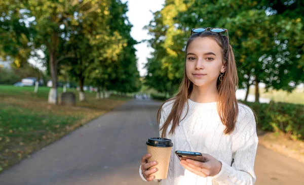 漂亮的少女女学生、暑期公园、手持智能手机、茶杯和咖啡。临时保暖的衣服毛衣太阳镜大学毕业后休息一下吧。积极放松的情绪. — 图库照片