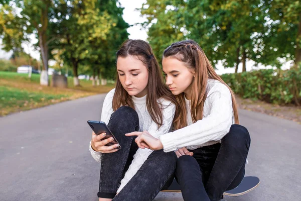 Girls girlfriends teenagers, summer in park, video on phone, rest after school. SMS on smartphone. Read write message, online social networks on Internet. Skateboard, trees road background.