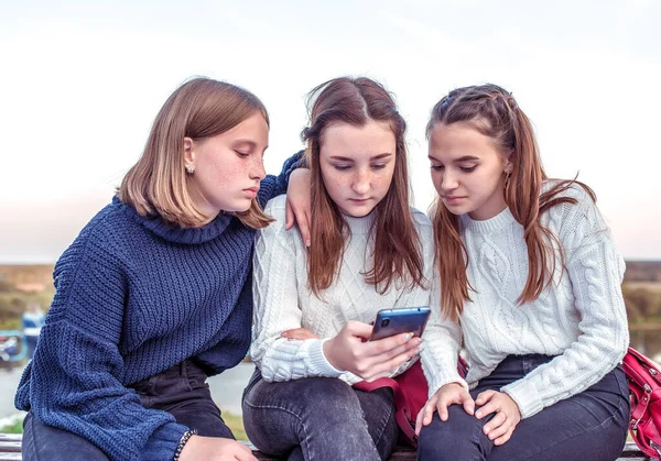 Tři dospívající školačky, na podzim na lavičce, sledují videa na smartphonu, internetovou síť v telefonu. Online aplikace. Teplé pletené svetry. Emoce relaxace po škole. — Stock fotografie