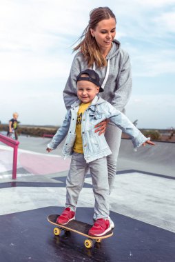Woman mom, holds little boy son 3-5 years old, learning to ride a skateboard, in the summer on the sports ground, emotions of happiness, fun, relaxation and pleasure. Casual wear.