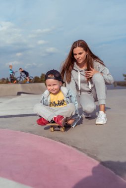 Woman mom, trains on skateboard little boy son 3-5 years old, learning to ride a skateboard, in the summer on sports ground, emotions of happiness, fun, relaxation and pleasure. Casual wear.