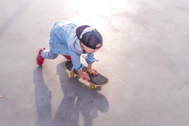 Little boy 3-5 years old, view from the top, in summer on sports ground, learning to ride a skateboard, casual wear, jeans, pants, a baseball cap. Free space for copy text.