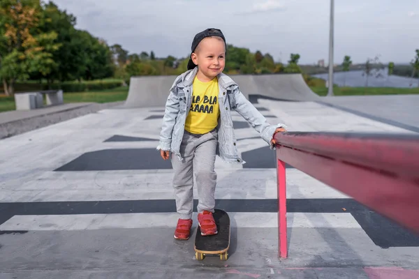 Little happy boy tersenyum, di musim panas di lapangan olahraga, belajar untuk naik skateboard, pakaian santai, celana jeans, celana, topi bisbol. Emosi relaksasi dan kenikmatan positif dan kesenangan. — Stok Foto