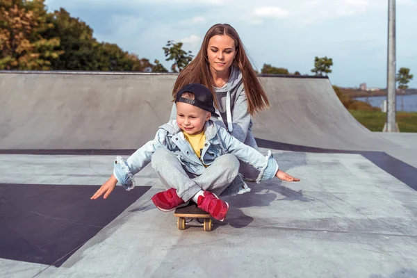 Ibu wanita, berseluncur di skateboard anak laki-laki berusia 3-5 tahun, belajar mengendarai skateboard, di musim panas di lapangan olahraga, emosi kebahagiaan, menyenangkan, relaksasi dan kesenangan. Pakaian kasual . — Stok Foto