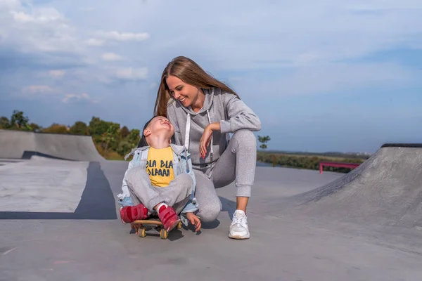 Ibu wanita senang tersenyum, menunggangi anak laki-laki berusia 3-5 tahun di skateboard, belajar mengendarai skateboard, di musim panas di lapangan olahraga, kebahagiaan emosi, kesenangan relaksasi yang menyenangkan. Pakaian kasual . — Stok Foto