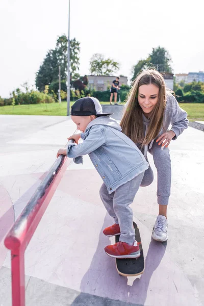 Ibu wanita membantu untuk skateboard anak laki-laki berusia 3-5 tahun, belajar untuk naik skateboard, musim panas di tanah olahraga, emosi kebahagiaan, menyenangkan, kesenangan relaksasi. Pakaian kasual. — Stok Foto