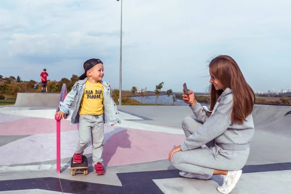 Woman mom photographs phone, little boy son 3-5 years old, learning to ride skateboard, summer on sports ground, emotions happiness, fun, relaxation pleasure. Record video on a smartphone. — Stok fotoğraf