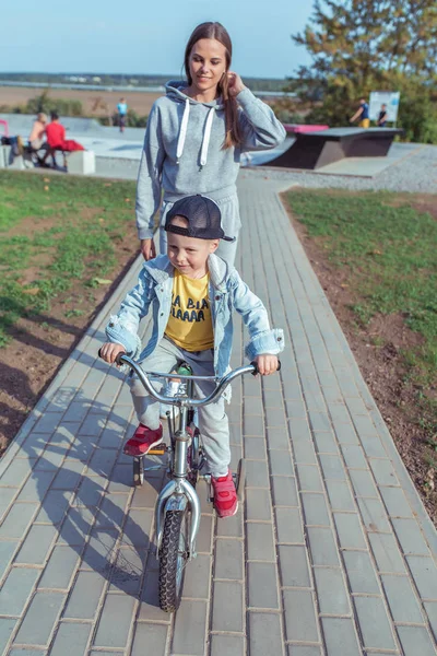 Ibu muda merawat anak kecil, berlatih dan belajar mengendarai sepeda, di musim panas di kota. Latar belakang jalan lapangan dan ubin. Emosi kebahagiaan adalah kesenangan dan sukacita. — Stok Foto