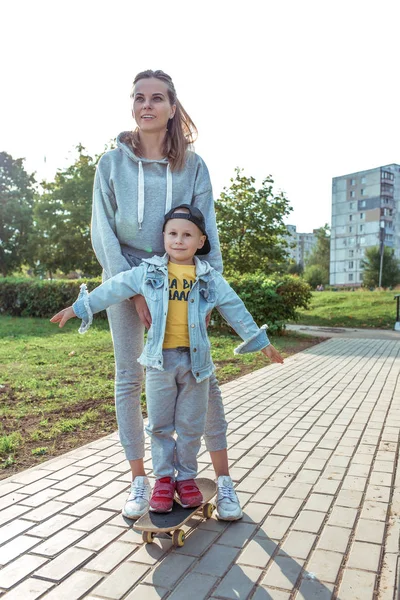 Familie tânără fericită, mamă femeie cu băiat copil, skateboarding, asistență parentală, parenting, primele lecții de skateboarding. În oraşul de vară. Emoțiile de râs sunt bucurii de distracție și plăcere. . — Fotografie, imagine de stoc