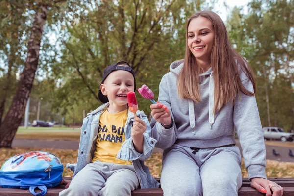 Woman mom with her son, little boy eats pink ice cream on stick. In summer city, casual clothes, happy rejoices, has fun delighted with delicious ice cream. Background trees are sitting on bench. — Stockfoto