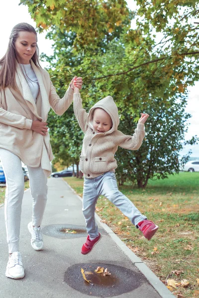 Keluarga muda, ibu wanita, berpegangan tangan, anak laki-laki kecil, musim gugur. Mereka berjalan di sepanjang jalan dan melompat melalui genangan air. Emosi menyenangkan tersenyum sukacita. Pakaian hangat dengan penutup kepala. Peduli terhadap pengasuhan anak. — Stok Foto