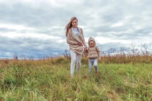 Ibu wanita berjalan dengan anaknya, hari musim gugur di taman, latar belakang lapangan, angin yang kuat, awan, ruang kosong untuk menyalin teks. Berjalan di pedesaan di alam. Pakaian hangat kasual . — Stok Foto