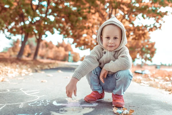 Liten glad pojke ler och sitter och vilar, ritar med färgade kritor på asfaltteckningar, kreativ konst och kreativitetskoncept. Bakgrundsträd i parken på hösten. Fritt utrymme för kopieringstext. — Stockfoto