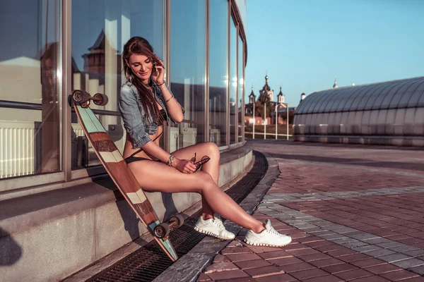 Beautiful stylish and fashionable girl summer in city calls by mobile phone, relaxes and sunbathes, longboard skateboard. Free and open space for text. Modern style of youth on vacation. — Stock Photo, Image