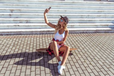 Güzel bir kız elinde cep telefonu, akıllı telefonunda selfie fotoğrafı, yazlık kıyafetler. Uzun tahta, paten, sosyal ağlar online uygulama. Duygular neşe ve rahatlama.