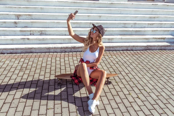 Schönes Mädchen mit Handy in der Hand, Selfie-Foto auf dem Smartphone, sommerliche Freizeitkleidung. Long Board, Skate, soziale Netzwerke Online-Anwendung. Emotionen erfreuen Spaß und Entspannung. — Stockfoto