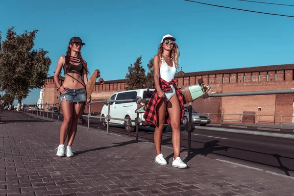 Duas meninas namoradas andando cidade verão, fundo é construção de carro de estrada, roupas casuais, mãos longboard skate. Caminhe no fim de semana, emoções relaxamento conforto prazer. Espaço livre para texto . — Fotografia de Stock