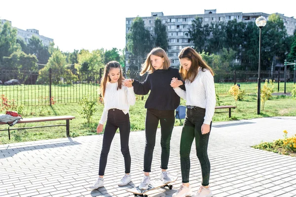 Drei Mädchen im Teenageralter fahren im Sommer in der Stadt Skateboard, Pause nach dem Schulwochenende. Lässig warme Kleidung Pullover Jeans. Emotionen der Freude Spaß an der Unterhaltung. Hintergrund Rasenbäume Gebäude. — Stockfoto