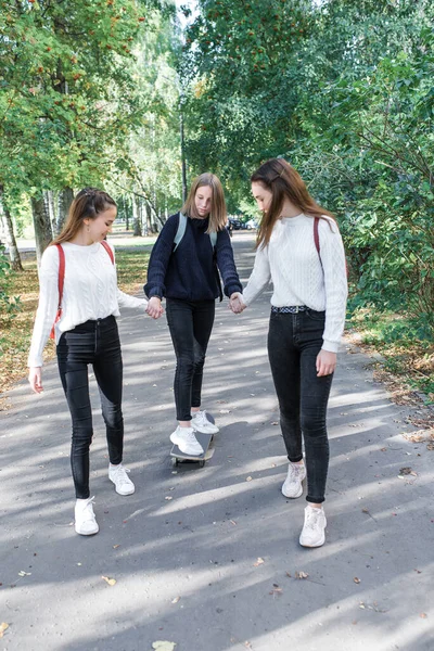 Drei Mädchen im Teenageralter fahren im Sommer in der Stadt Skateboard, Pause nach dem Schulwochenende. Lässig warme Kleidung Pullover Jeans. Emotionen der Freude Spaß an der Unterhaltung. Hintergrund Rasenbäume . — Stockfoto