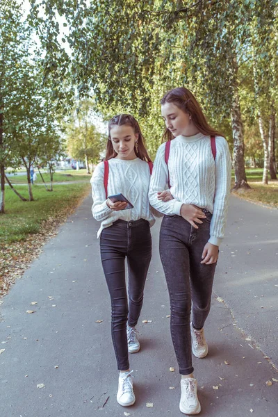 2人の女子学生、手で歩いて、学校の後に家に戻る。夏の都市では、背景の道路の木、手のスマートフォン。後ろのバックパック。普段着のセータージーンズ。ソーシャルネットワークオンライン. — ストック写真