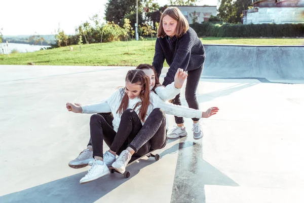 3 gadis sekolah naik skateboard, beristirahat setelah sekolah institut akhir pekan kuliah. Kota musim panas, lapangan olahraga. Emosi kebahagiaan, menyenangkan, sukacita, tawa, senyum, hiburan. Casual memakai jeans sweater . — Stok Foto