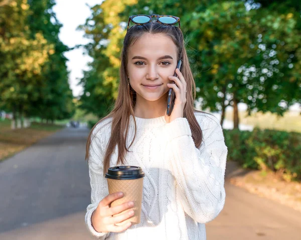 Счастливая девочка радуется, подросток 12 лет, звонит рука смартфона, чашка кофе, слушает голосовое сообщение, онлайн-приложение в Интернете. Социальные сети. В летнем парке. Отдых в выходные . — стоковое фото