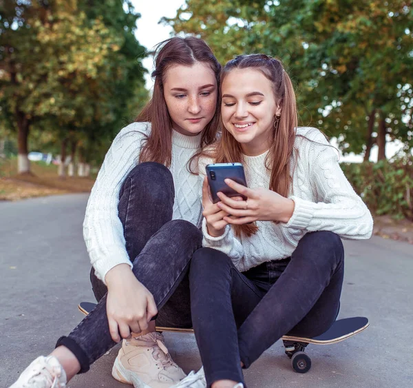 Dvě dospívající dívky dospívající, v letním parku, sedí na skateboardu, sledují videa na smartphonu, on-line aplikace na internetu. Emoce štěstí zábavný smích radosti. Vtipné video na telefonu. — Stock fotografie