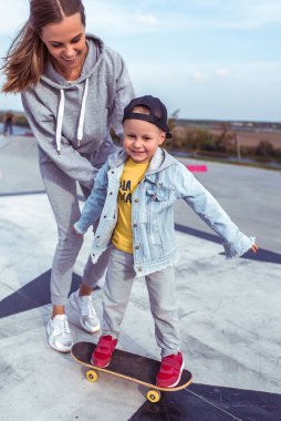 Mutlu aile, kadın anne, kaykaydaki kaykaycı çocuk, spor fitness eğitimine yardım. Mutluluk duyguları rahatlama ve neşe, gülümseme ve kahkaha. Yaz sporları sahası.