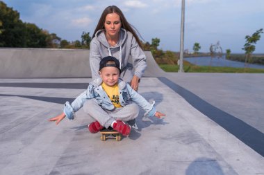 Aile kadını, kaykaydaki çocuğun oğlu anne, spor antrenmanlarına yardım ediyor. Mutluluk duyguları, rahatlama sevinci, gülümseme kahkahası. Yaz sporları sahası. Metni kopyalamak için boş alan.