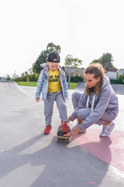 Kvinna mamma med en liten pojke son, utbildning på en skateboard, hjälp och stöd i idrott och fitness. Vårdande föräldraskap och stöd. På sommaren på idrottsplats. — Stockfoto