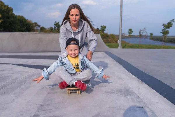 Wanita keluarga ibu dari anak laki-laki di skateboard, membantu mendukung pelatihan kebugaran olahraga. Emosi kebahagiaan, kesenangan relaksasi sukacita, senyum tawa. Lapangan olahraga musim panas. Ruang kosong untuk menyalin teks . — Stok Foto