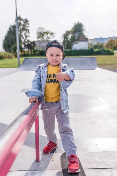 Niño pequeño de 4-5 años, monta en un monopatín, en verano en la ciudad en el campo de deportes. Ropa casual. Emociones de positivo y divertido, una sonrisa y alegría en la cara. Súper gesto de mano, dedo arriba . —  Fotos de Stock