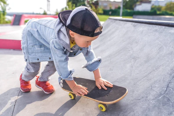 Anak kecil berusia 4-5 tahun, naik skateboard, di musim panas di kota di lapangan olahraga duduk. Pakaian kasual denim dan topi baseball. Ruang kosong untuk menyalin teks. Pelajaran pelatihan pertama . — Stok Foto