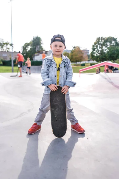 Anak kecil berusia 4-5 tahun, berdiri dengan skateboard, di musim panas di kota di lapangan olahraga duduk. Pakaian kasual denim dan topi baseball. Emosi senyum positif dan sukacita . — Stok Foto