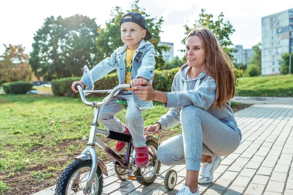Keluarga muda, ibu, anak laki-laki berusia 3-5 tahun, musim panas di kota. Belajar mengendarai sepeda, latihan keseimbangan, membantu orang tua membesarkan anak. Emosi yang positif dan kebahagiaan . — Stok Foto