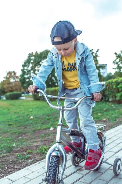 Anak kecil berusia 3-5 tahun, di kota musim panas. Kereta belajar untuk menjaga keseimbangan pada sepeda, naik pedal. Di kota musim panas, kepentingan emosi adalah kenyamanan kebahagiaan. Pakaian santai, topi baseball denim, sepatu olahraga . — Stok Foto