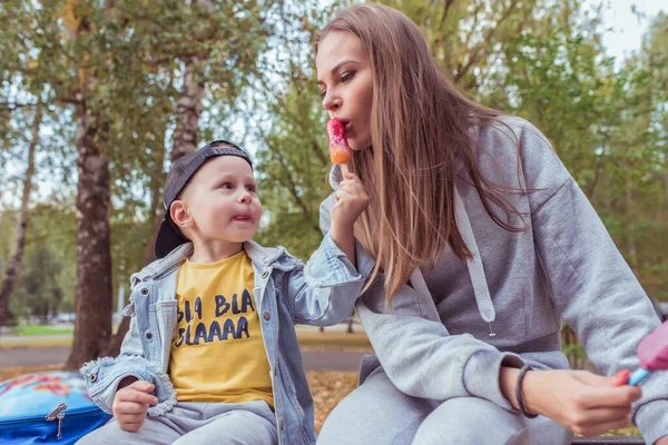 Kvinna mamma med en liten pojke son, äta glass på en pinne. Sitter på en bänk. Bakgrundsträd, sommar i park. Vårdande föräldraskap och stöd. Snack för lunch läckra sötsaker i naturen — Stockfoto