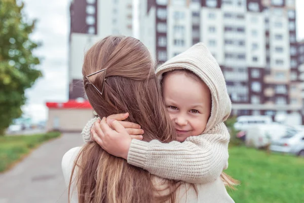 Küçük çocuk şehirde sonbaharda kadın anneye sarılıyor. Kapüşonlu sıcak giysili bej kazak. Mutluluk duyguları gülümseme ve zevk aşkı. Sarıl bana. Çocuğun bakımı ve ebeveynliği. — Stok fotoğraf