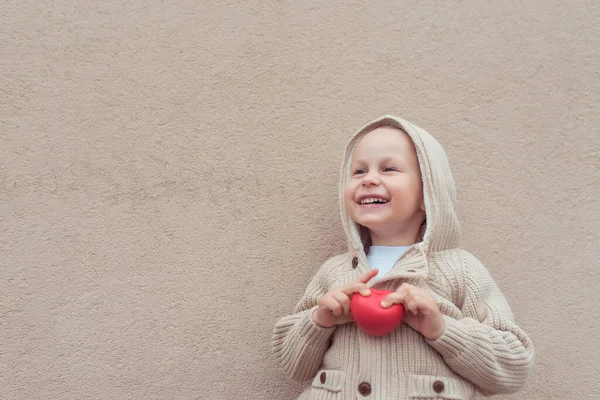 Feliz menino alegre de 3-5 anos de idade segurando coração vermelho em suas mãos brinquedo, parede de fundo, alegre em positivo. Espaço livre para texto de cópia, banner para tagline. Emoções diversão alegria celebração . — Fotografia de Stock