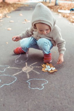 Kapüşonlu bej kazaklı küçük bir çocuk, yazın yaz günü, asfalta renkli tebeşirlerle güneş çizer. Yaratıcı fikir ve yaratıcılık.